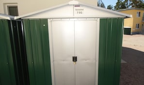 A green metal storage shed with a white door, labeled "Hasenwinkel 7 EG".
