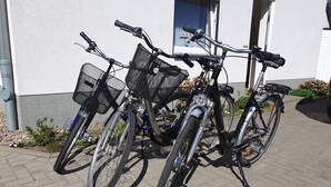Two bicycles parked on a paved area in front of a building with a white exterior and a window with a blue curtain.