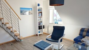 A modern living room with a staircase, a bookshelf, a blue armchair, and a glass coffee table.