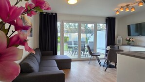 Een moderne woonkamer met een grijze bank, een eethoek met een tafel en stoelen, en een keukenhoek met een televisie en hanglampen.