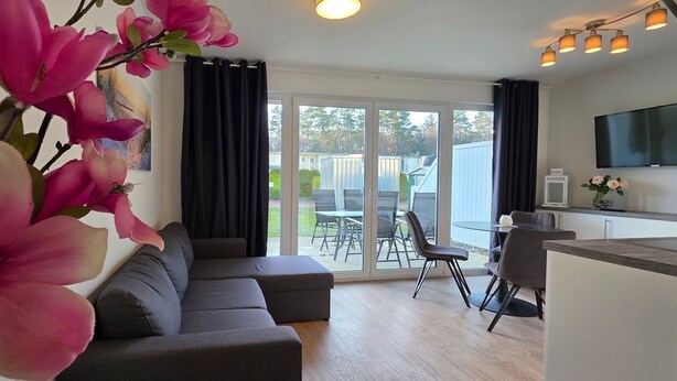 A modern living room with a grey sofa, a dining area with a table and chairs, and a kitchen area with a TV and pendant lights. - Apartment KWV 1 ground floor - 2-room apartment for 3 pers. on the ground floor and garden (approx. 30 m²) (Graal-Müritz)