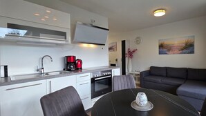 A compact kitchen with modern appliances, a dining area with a table and chairs, a sofa, and a painting on the wall. - Apartment KWV 1 ground floor - 2-room apartment for 3 pers. on the ground floor and garden (approx. 30 m²) (Graal-Müritz)