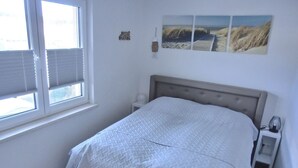 A bedroom with a bed, a nightstand, a bench, a window with blinds, and a wall-mounted lamp. - Apartment KWi 77 OG "Windflüchter" - 2-room apartment for 3 persons on the upper floor with (Graal-Müritz)