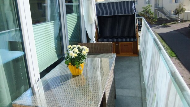 A balcony with a glass table, a wicker chair, and a yellow vase with flowers. - Apartment KWi 77 OG "Windflüchter" - 2-room apartment for 3 persons on the upper floor with (Graal-Müritz)