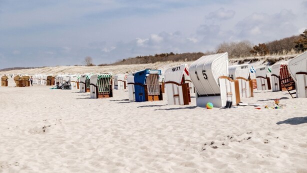 Una fila di casette sulla spiaggia, con numeri e la scritta "WITT", disposte su una spiaggia sabbiosa sotto un cielo sereno.