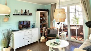 Una sala de estar moderna con un mueble blanco, un televisor, una silla de mimbre, una mesa redonda y un ventanal con cortinas.