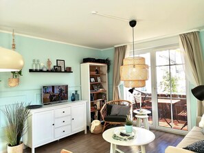 Un salon moderne avec un meuble blanc, un téléviseur, un fauteuil en osier, une table ronde et une grande fenêtre avec des rideaux.