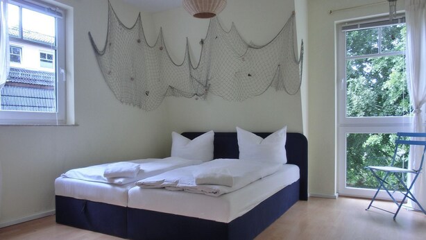 Una cama bien hecha en una habitación con un ventanal grande, una lámpara colgante y una silla plegable azul.
