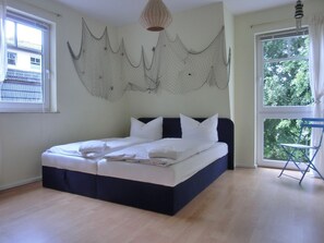 Un lit bien fait dans une pièce dotée d’une grande fenêtre, d’un luminaire suspendu et d’une chaise pliante bleue.