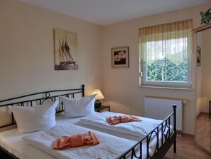 A double bed with white linens and two pink towels, a nightstand with a lamp, a window with a view of greenery, and a framed picture on the wall.