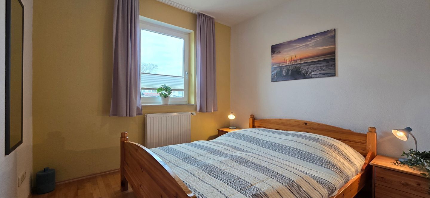 Quarto com cama de madeira, janela com cortinas, radiador, luminária de mesa e pintura na parede.
