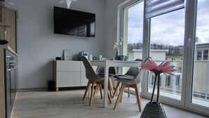Et moderne køkken med et fladskærms-tv, to stole, et spisebord og et stort vindue med udsigt til bygninger.