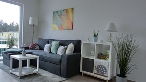 En moderne stue med en grå sofa, et lille hvidt sofabord, en bogreol og et stort vindue.