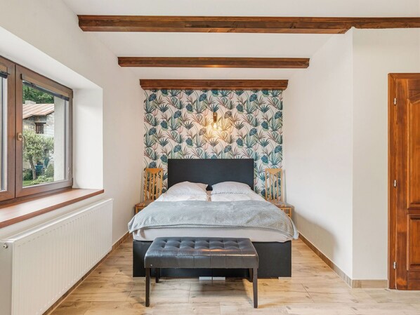 A bedroom with a bed, bench, wooden beams, radiator, and a window.