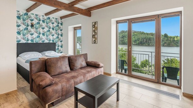 Une chambre avec un lit, un canapé marron, une petite table, un balcon donnant sur un lac et un papier peint à motifs.