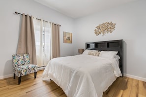 Une chambre avec un lit, une chaise, un tableau décoratif au mur et une fenêtre avec des rideaux.