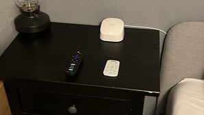 Una mesita de noche negra con una lámpara, un control remoto y un dispositivo electrónico blanco.
