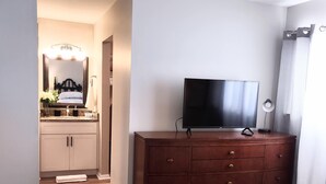A bedroom with a wooden dresser, a flat-screen TV, a bed with a patterned bedspread, and a bathroom visible through an open door. - Idyllic Central Raleigh condo-3br/2bath (Raleigh)