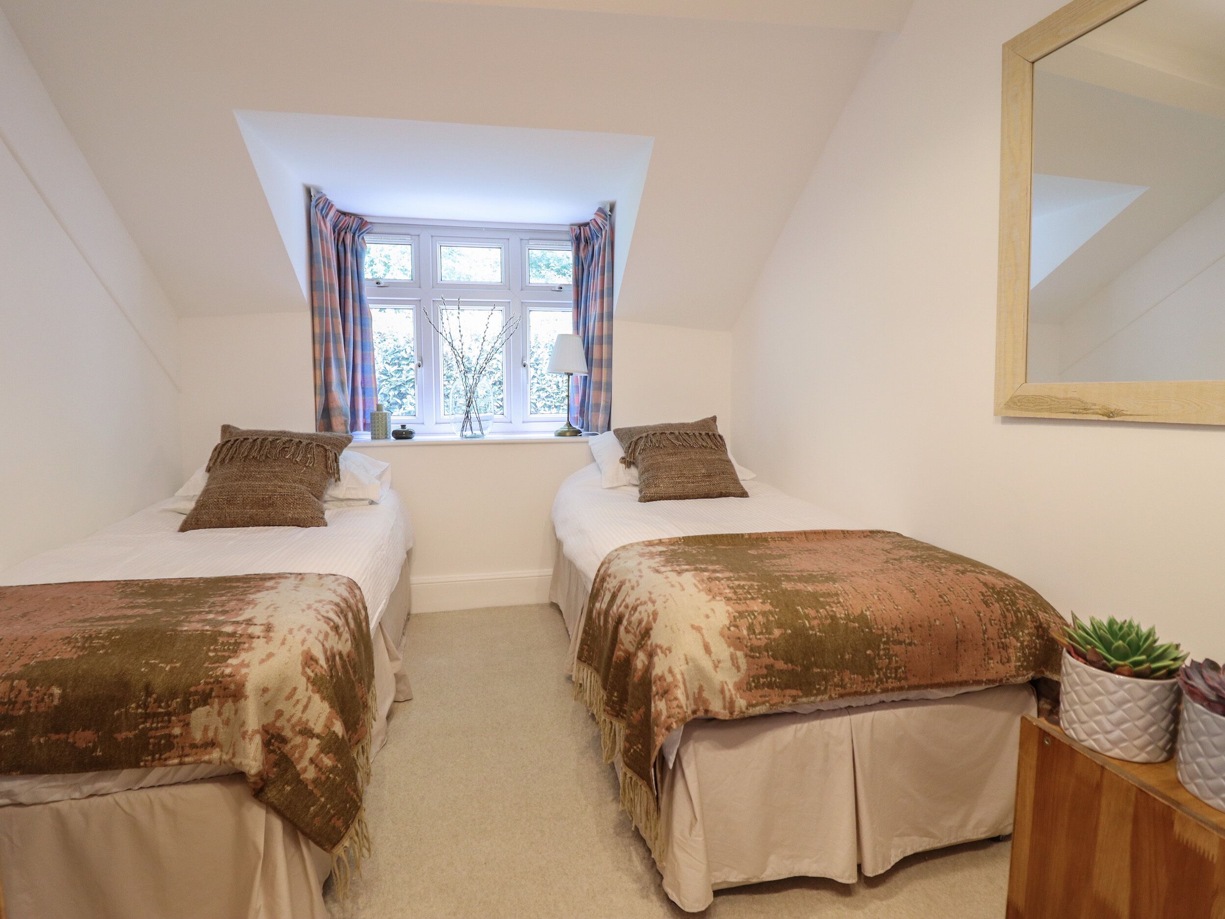 A bedroom with two beds, a window with curtains, a mirror, and a small plant.
