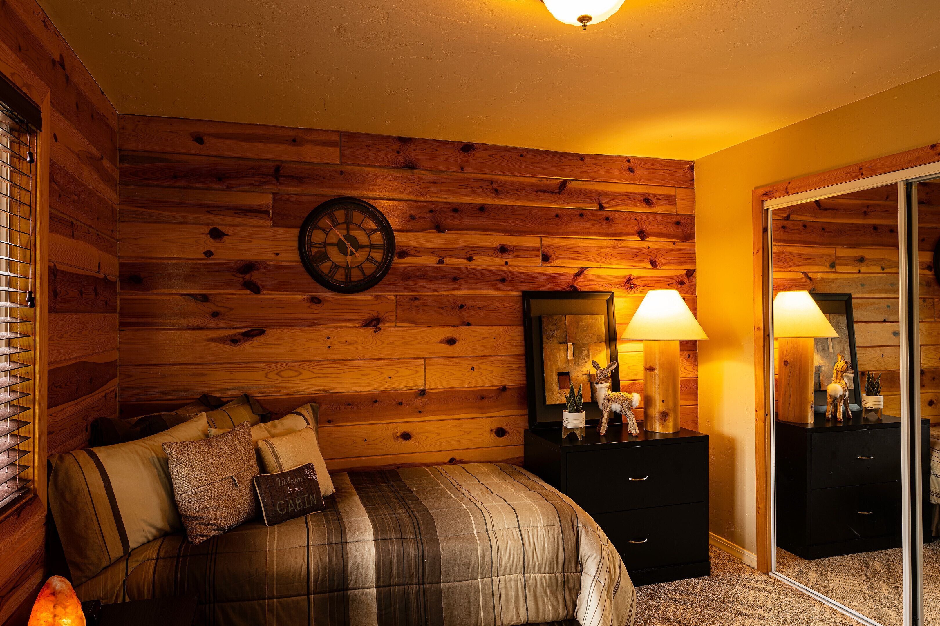 A cozy bedroom with a wooden wall, a bed with striped bedding, a nightstand with a lamp, and a mirror.