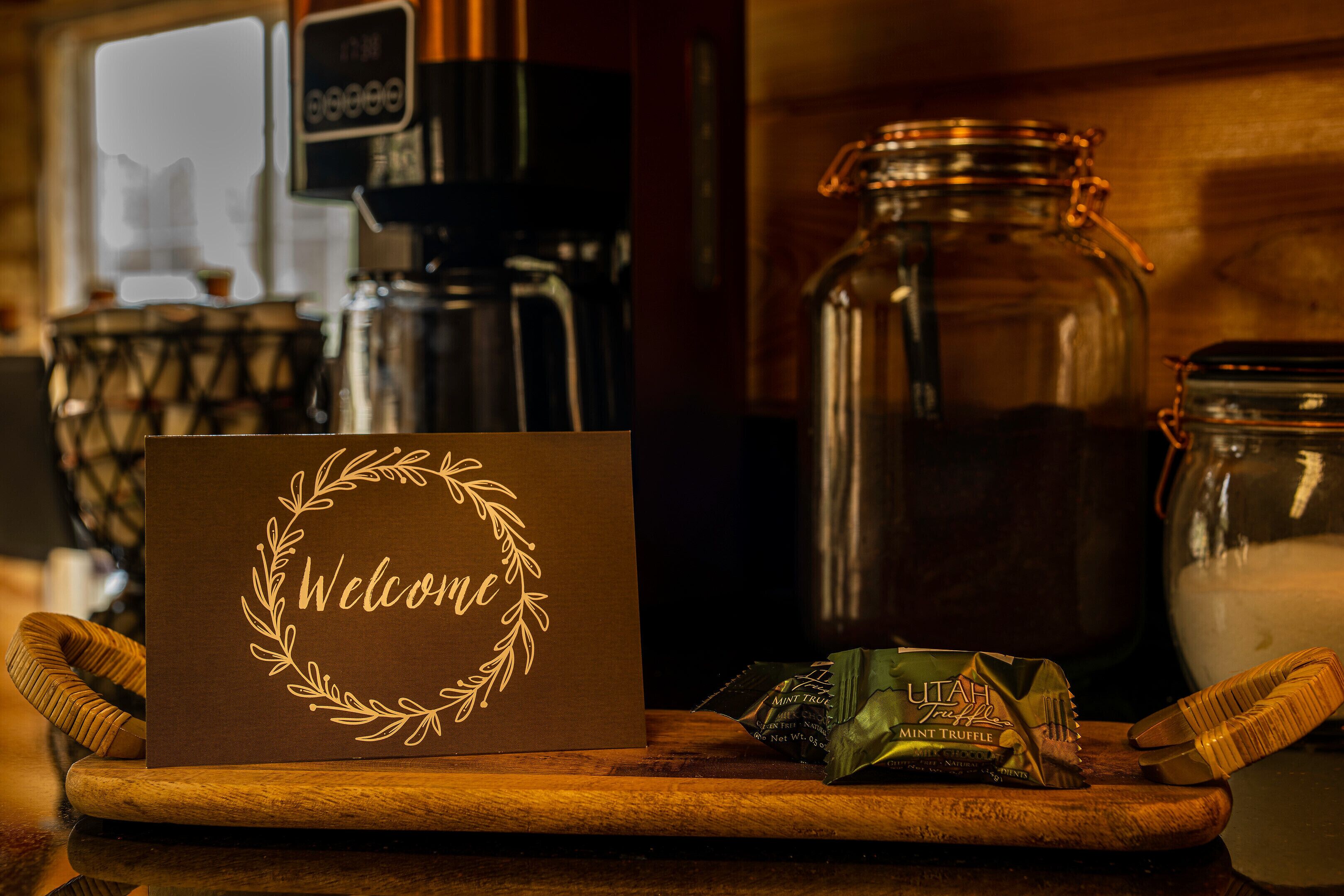 Una tarjeta de bienvenida con diseño de laurel sobre una mesa de madera, una cafetera, un tarro de cristal y un paquete de trufas de menta.