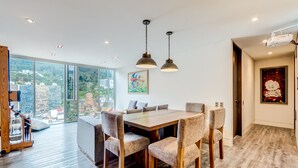 A modern dining area with a wooden table and chairs, large windows offering a city view, and pendant lighting.
