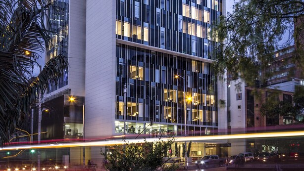 A modern building with a grid of windows, a paved plaza with a tree, and a street with moving lights.