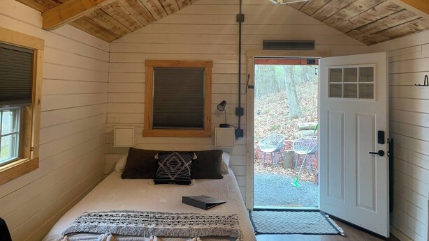 A cozy bedroom with a wooden ceiling, a bed with a patterned throw, a window with blinds, and a door leading outside. - Shawnee Tiny Cabin near Ferne Clyffe with Hot Tub (Goreville)