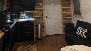A cozy cabin interior with a wooden ceiling, a central light fixture, a kitchen area with appliances, a door, and a black couch with a patterned pillow. - Shawnee Tiny Cabin near Ferne Clyffe with Hot Tub (Goreville)