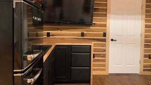 A compact kitchen with a built-in refrigerator, a stove, a microwave, and a flat-screen TV mounted on the wall. - Shawnee Tiny Cabin near Ferne Clyffe with Hot Tub (Goreville)