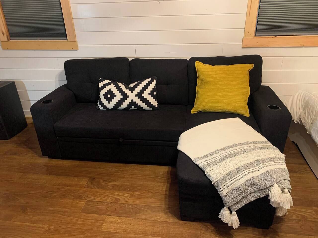 Un canapé noir agrémenté d’un coussin à motifs et d’un coussin jaune, une couverture blanche et un plancher en bois.