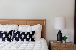 Un lit bien fait, avec des oreillers blancs et bleu marine, une tête de lit en bois, une lampe de chevet, une petite plante en pot et une télécommande posée sur une table de chevet en bois.