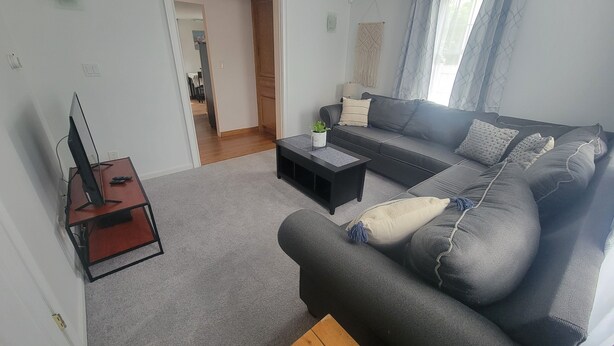 Ett vardagsrum med en mörk soffa, ett soffbord i trä, en tv på ett tv-bord och en dörr som leder till ett annat rum.