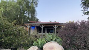 A rustic cabin with a red roof, surrounded by large rocks and various plants. - PCH Bunkhouse (San Luis Obispo)