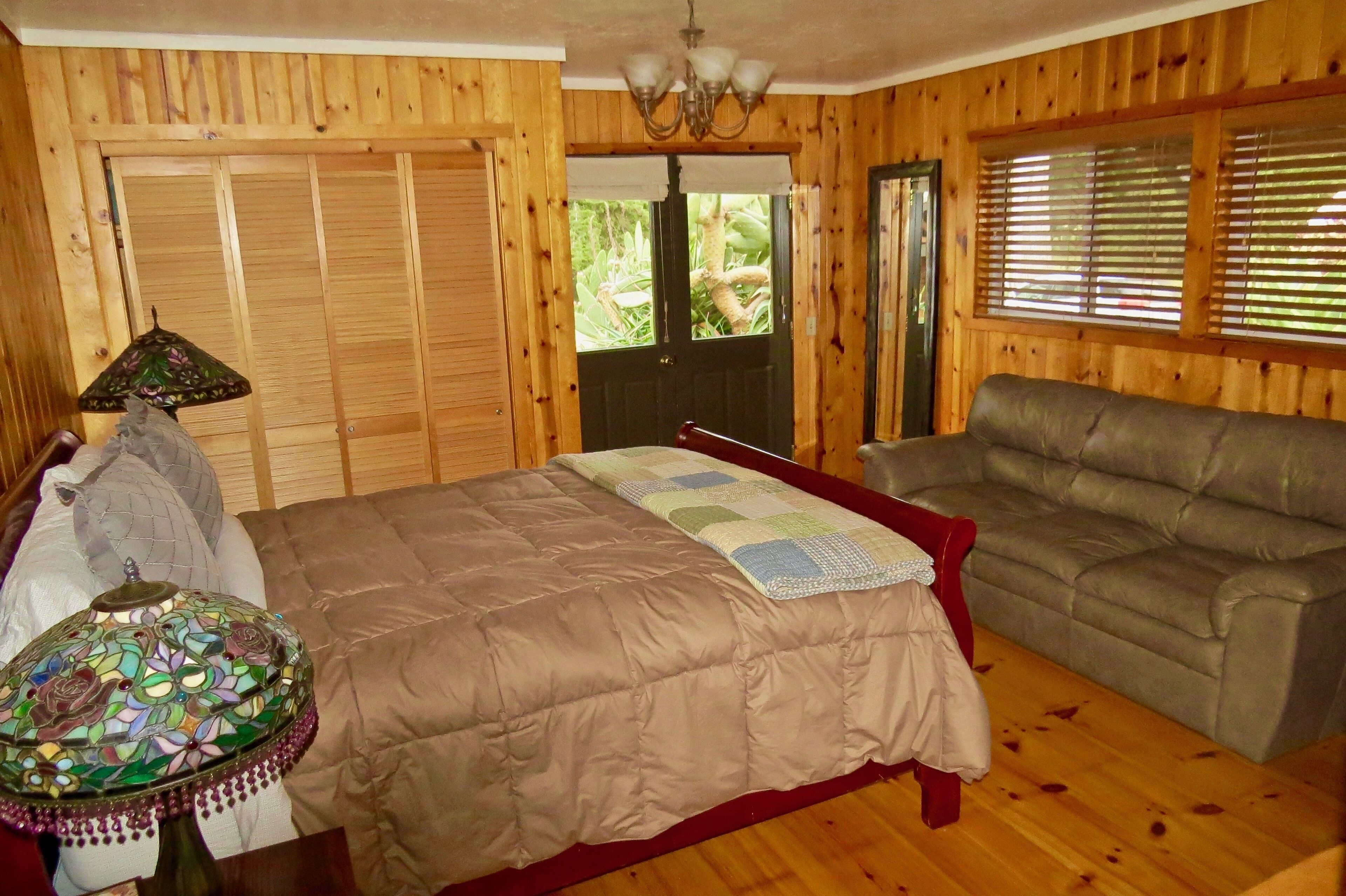 Una habitación con paneles de madera, una cama, un sofá, una mesa con lámpara y una ventana con persianas.
