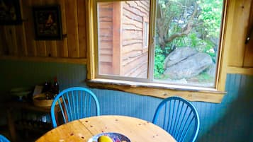 Interior de uma cabana de madeira com uma mesa de jantar, cadeiras azuis e uma janela com vista para um jardim com uma grande rocha.