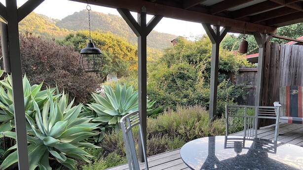 A covered outdoor patio with a round table and chairs, surrounded by various plants and a view of a mountainous landscape. - PCH Bunkhouse (San Luis Obispo)