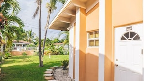A house with a white door, a covered porch, and a garden with palm trees and a white fence.