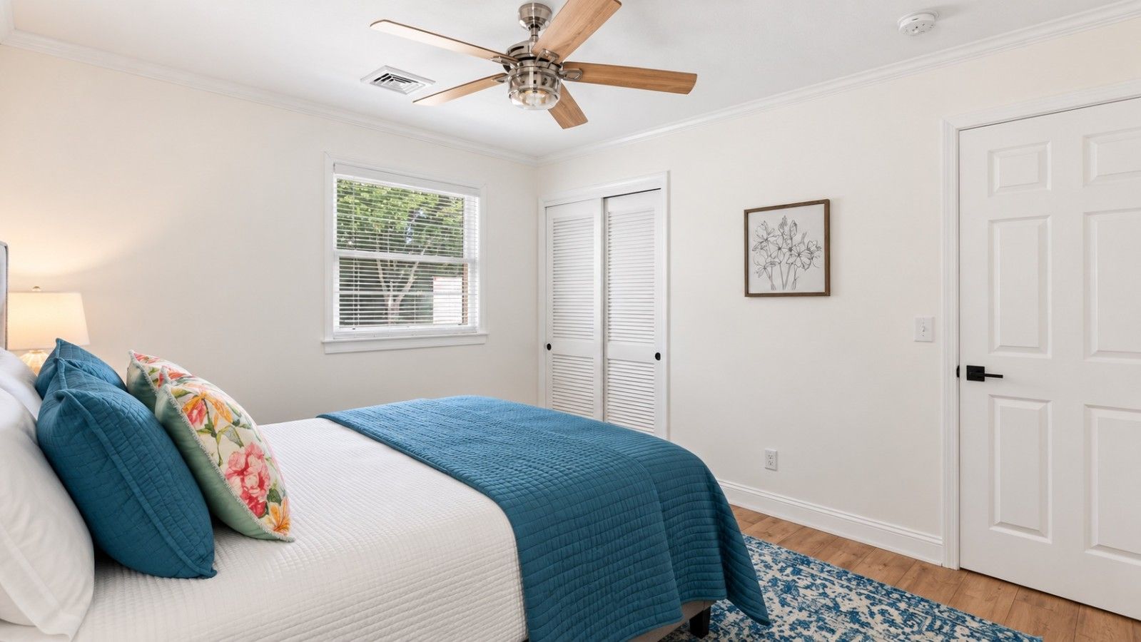 Une chambre à coucher comprenant un lit, un ventilateur de plafond, une fenêtre avec des stores, une porte et un tableau encadré accroché au mur.