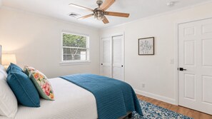 Un dormitorio con cama, ventilador de techo, ventana con persianas, puerta y un cuadro en la pared.