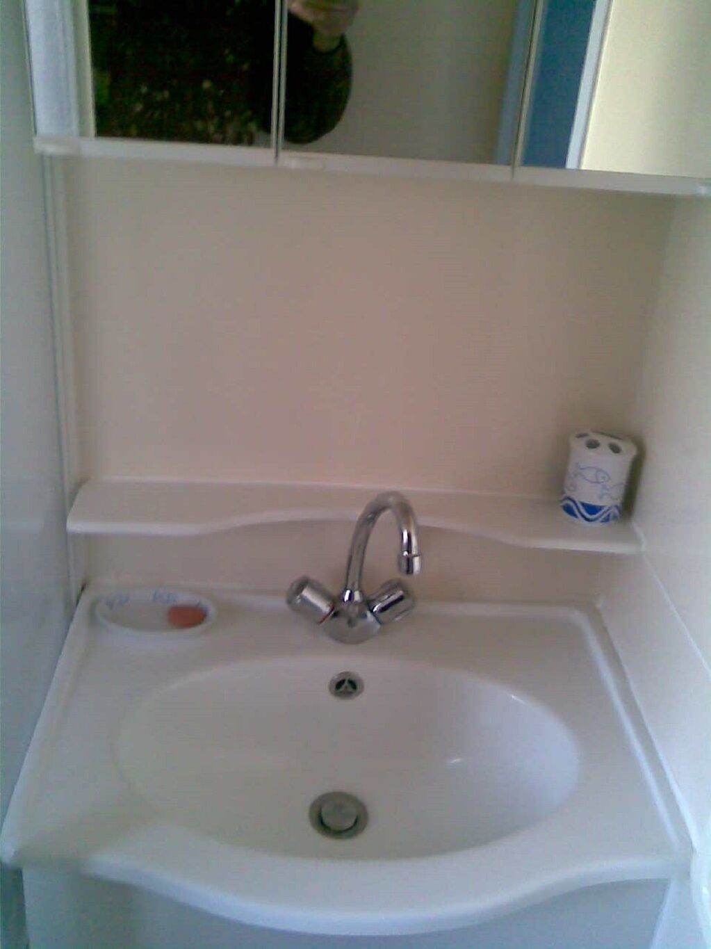 A white bathroom sink with a chrome faucet, a soap dish, and a toilet paper holder on a white shelf.
