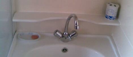 A white bathroom sink with a chrome faucet, a soap dish, and a toilet paper holder on a white shelf.