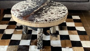 Un poggiapiedi rotondo rivestito in pelle con decorazioni a macchia e un bordo intrecciato, posto su un tappeto a scacchi.