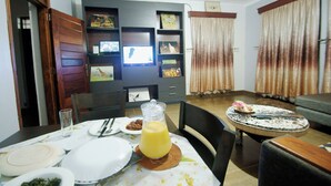 Zona pranzo con tavolo apparecchiato per due, televisione a parete e libreria con diversi oggetti.
