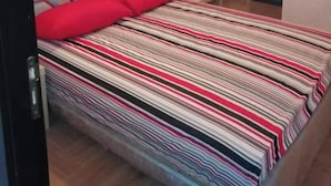 Makuuhuone, jossa on sänky, punainen tyyny, raidallinen päiväpeite, yöpöytä ja riippuvalaisin.