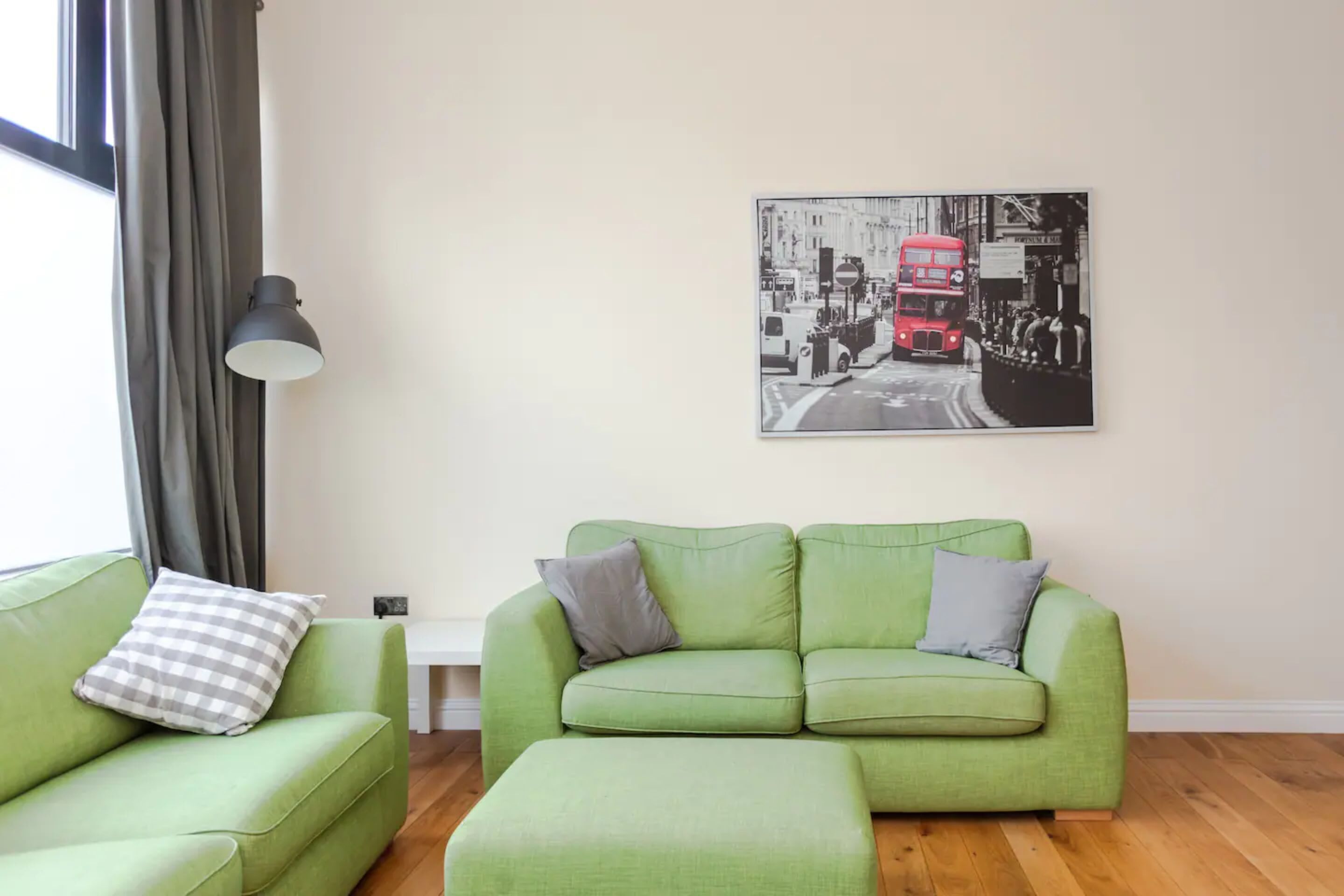 A living room with a green sofa and ottoman, a black lamp, and a black and white photograph of a red double-decker bus on the wall.