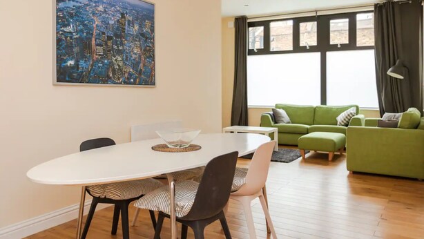 A modern living room with a dining table, green sofa, and a large window with curtains. - Stylish 2 Bedroom Apartment in Greenwich (London)