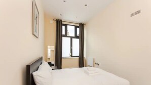 A neatly made bed in a hotel room with a window, a framed picture on the wall, and a small table with a chair. - Stylish 2 Bedroom Apartment in Greenwich (London)
