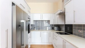 A modern kitchen with white cabinets, a stainless steel refrigerator, a microwave, and a black tiled backsplash. - Stylish 2 Bedroom Apartment in Greenwich (London)