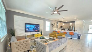 A modern living room with a TV, sofa, and chairs, a kitchen in the background, and a ceiling fan. - Disney theme park 10 mmins/Storey Lake 9 Beds /Lake View Single House (kissimmee)